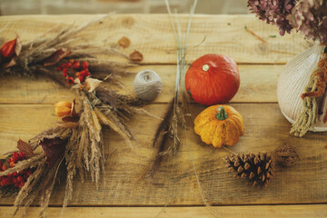 Stylish autumn wreath on rustic table. Fall decor and arrangement in farmhouse. Rustic autumn wreath with dried grass, berries, herbs on wooden table with pumpkins and pine cone.