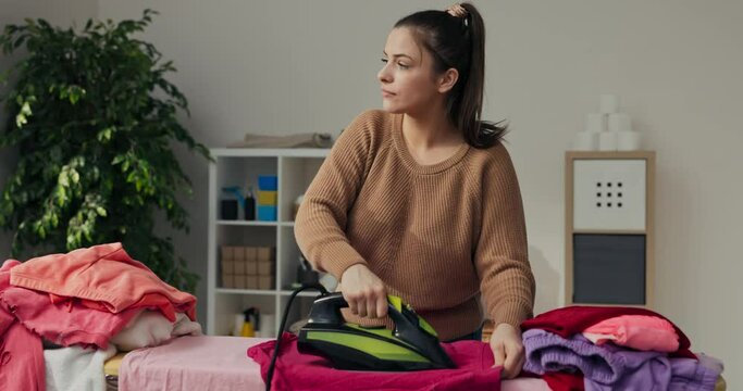 Busy mother stands in the room and irons clothes. There are clothes on ironing board and she is ironing pink T-shirt with green iron. Woman looked out the window, rejoiced at sunny weather and smiled.