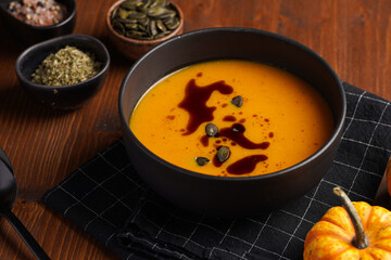 One deep black bowl with an orange pumpkin cream soup with pumpkin seed oil, two black spoons on a wooden surface
