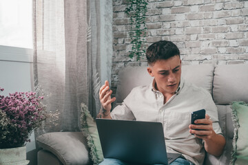 Angry young man using computer pc at home sitting on the sofa having problems with his phone. Mad...