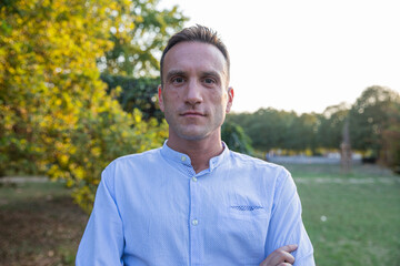 Portrait of a man in his 30s standing still in the park with crossed arms looking at the camera.