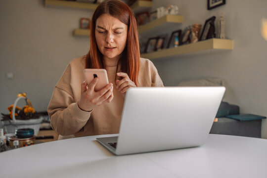 Unhappy young woman looking at smartphone screen, irritated by bad gadget work, low internet connection, working remotely at home. Stressed attractive lady annoyed by hard work task or system crash. 