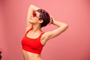 Fashion portrait of a woman with a short haircut in colored sunglasses with unusual accessories with earrings smiling on a pink bright background