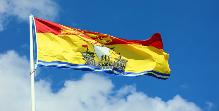 The flag of New Brunswick consists of a golden lion passant on a red field in the upper third and a gold field defaced with a lymphad on top of blue and white wavy lines in the bottom two-thirds.