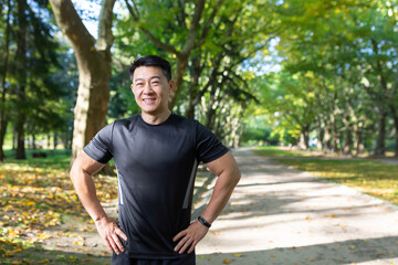 Portrait of Asian athlete in autumn park on sunny day, man in sportswear smiling and looking at camera, fitness instructor before training.