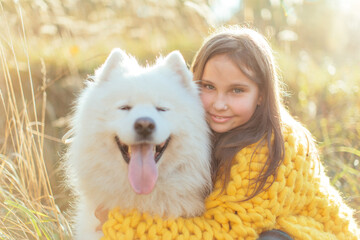 A little brunette girl in a knitted yellow coat with her white fluffy dog lies on autumn leaves in the park. Friendship and harmony in the family. Happy childhood.