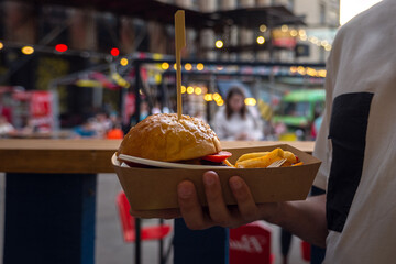 Man hands holding street food burger with french fries on craft paper. Street fast food. festival background