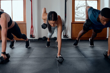 Three multiracial young adults in a row doing plank exercises with weights at the gym
