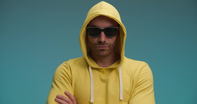 casual young man with yellow sweatshirt nodding, taking sunglasses off and making a funny face on blue background in studio