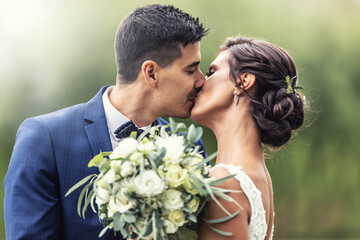 Newlyweds kiss outdoors while holding a wedding bouquet of white flowers in front of them