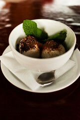 Green mint leaves in a bowl of vanilla ice cream with chocolate chips. A plate with garnished with grated chocolate ice cream on a brown wooden table. Summer desserts for a day off. Sweets and goodies