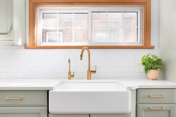 A kitchen sink detail shot with a gold faucet, farmhouse sink, subway tile backsplash, and a light green cabinet.