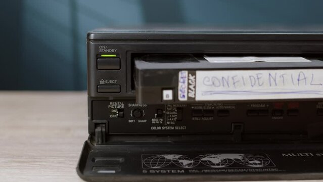 man inserts old black cassette tape into VHS player labeled CONFIDENTIAL. old vintage cassette VCR player is on table, player for viewing information on magnetic tape.