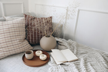Breakfast in bed, sofa stationery mockup scene. Cup of coffee, pumpkins. Open book. Cup of coffee on tray. Dry grass bouquet in ball shaped vase on linen throw. Lifestyle concept. Checkered cushions.