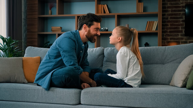 Playful caucasian young family daddy and small daughter sitting comfortable on couch loving cute girl touching father nose funny joke playing game with parent having fun spending free time together