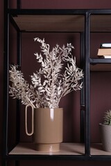 Stylish ceramic vase with dry plants on shelf near brown wall