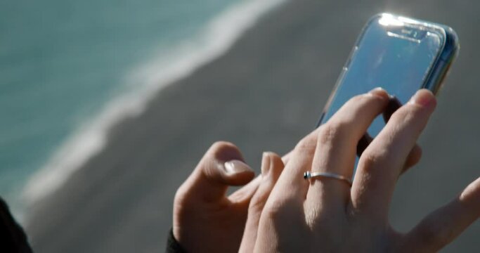 close up shot of a girl holding her phone and swipping with a black sand beach in the background