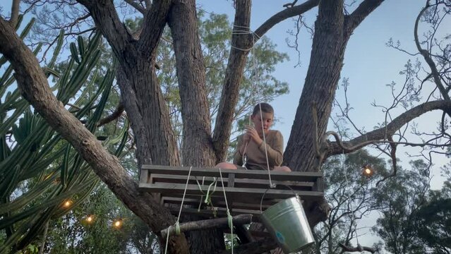 A curious boy pulls a bucket up to a simple DIY wooden pallet treehouse