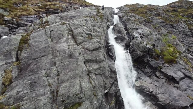Man flying drone from steep wilderness cliff with waterfall in Norway - Aerial flying upp mountainside and passing person before revealing full mountain landscape Storfjellet in Stamneshella