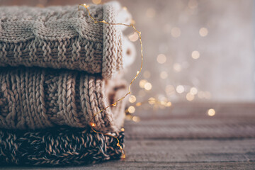 Stack of cozy winter or autumn knitted sweaters and garland lights on wooden background. Warm Cozy Concept