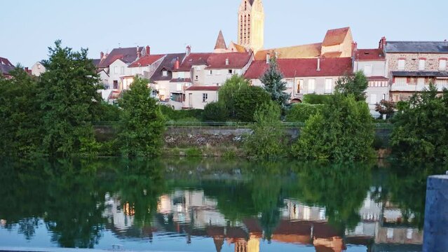 Scenic view of small French town of Dormans on bank of Marne river overlooking illuminated tower of Saint-Hippolyte church rising above houses 