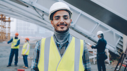 portrait of young Middle Eastern builder wearing uniform standing in construction site smiling and...