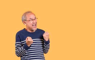 Yes! Portrait of Asian senior man wearing glasses standing raising his fists and excited facial expression isolated on yellow background.