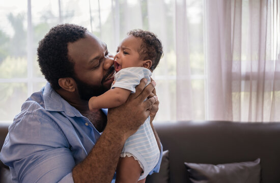 Happy African father man sitting holding and kissing newborn baby son on a sofa in the living room at home.