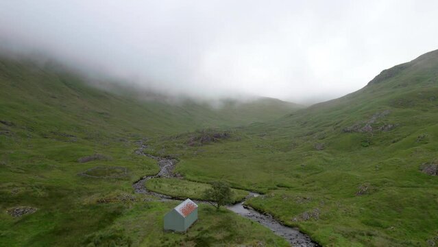 Aerial drone footage rising slowly up into atmospheric, low hanging cloud in a Scottish glen looking towards an old agricultural shed and a river as it winds its way up the glen. Scotland.