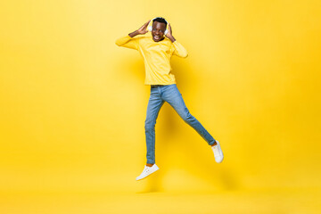 Fototapeta na wymiar Portrait of happy young African man wearing headphones listening to music and jumping in studio yellow color isolated background