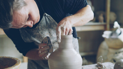 Close-up shot of skillful young potter molding ceramic vase from clay on throwing wheel while working in potter's workshop. Shelves with handmade eathenware in background.