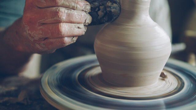 Elderly bearded craftsman is creating ceramic jar on potter's wheel using professional tools. Workplace with handmade pots, beautiful vases and figures in background.