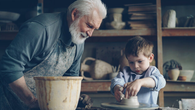 Concentrated young boy is molding clay into ceramic pot on spinning throwing wheel and his loving experienced grandfather is talking to him. Pottery and family tradition concept.