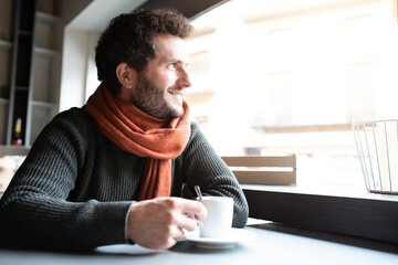 Pensive, happy young man drinking coffee in cafeteria looking out the window, enjoying early morning. Copy space. - Powered by Adobe