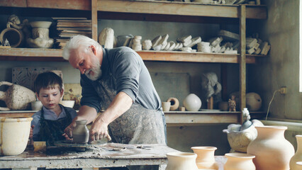 Professional potter is cutting ceramic pot from throwing wheel and his helpful little grandchild is bringing it to work table. Family members working together concept. - obrazy, fototapety, plakaty