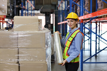 Caucasian warehouse worker woman with hardhat and reflective jackets wrapping boxes in stretch film...
