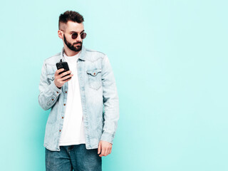 Handsome smiling model.Sexy stylish man dressed in jacket and jeans. Fashion hipster male posing near blue wall in studio. Holding smartphone. Looking at cellphone screen. Using apps