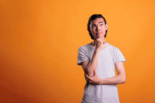 Pensive Asian Teenager Thinking, Making Choice, Touching Chin With Thoughtful Facial Expression, Idea Generation. Puzzled Young Man Looking Upwards, Medium Shot On Orange Background