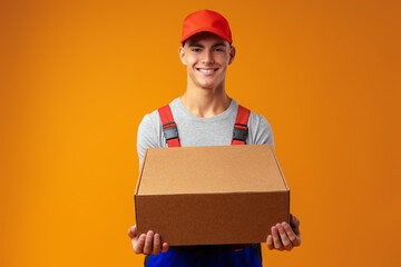 Young courier in uniform with box on yellow background in studio - obrazy, fototapety, plakaty