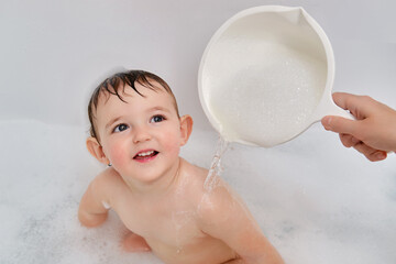 A mother washes a child sitting in a white tub with soap suds. Mom s hand waters the baby from a...