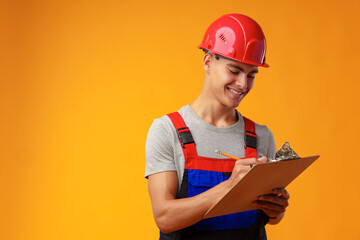 Young construction worker wearing hardhat and holding a clipboard on yellow background in studio