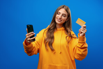 Portrait of a young blonde woman showing plastic credit card while holding mobile phone over blue background