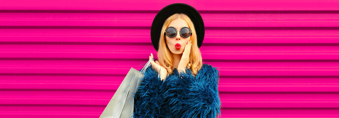 Portrait of beautiful stylish woman with shopping bags and blowing her lips sends air kiss with lipstick wearing blue fur coat, black round hat and sunglasses posing on pink background