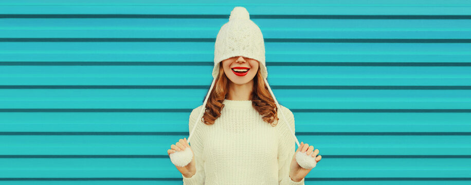Winter portrait of happy cheerful laughing young woman having fun wearing white knitted sweater, hat with pom pom on blue background