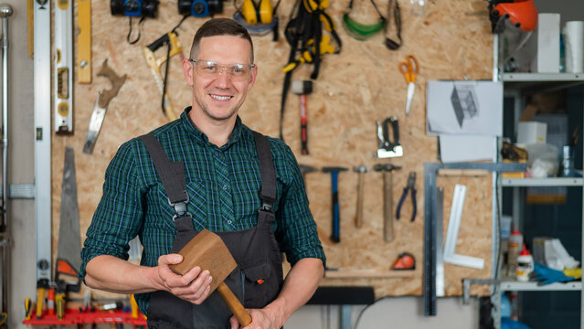 Portrait of a carpenter in goggles and overalls holding a wooden hammer in the workshop against the background of a wall with tools.