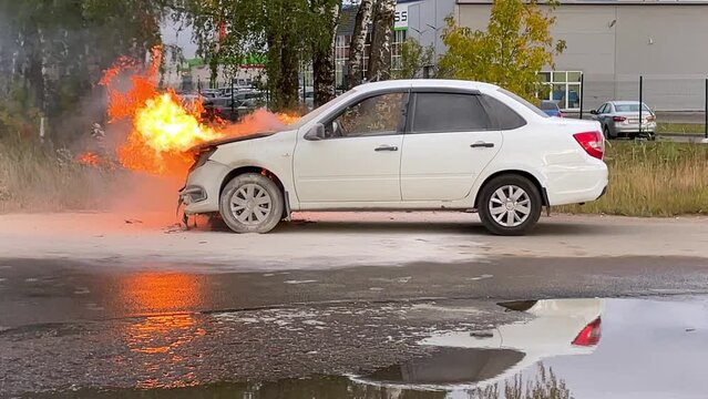 Accident. A white car in an industrial area is on fire. The engine compartment is on fire