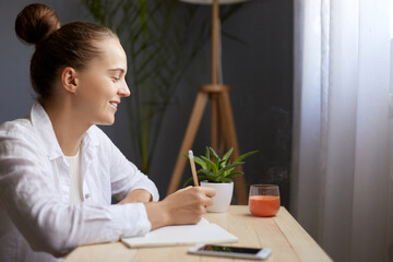 Profile portrait of smiling beautiful woman writing notes in paper notebook, thinking about something pleasant, dreaming, sitting at the table by the window.