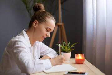 Side view portrait of dark haired attractive woman writing notes in paper notebook, sitting at the table by the window, making to do list or writes a composition.