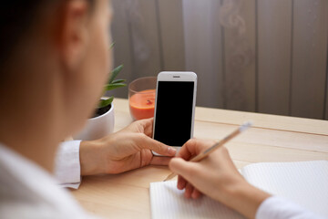 Portrait of unknown anonymous woman posing backwards and holding smart phone with empty display for advertisement, writing something in paper organizer.