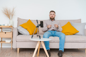 Smiling bearded guy filming video using smartphone on tripod online while sitting on sofa in living...